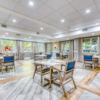 A modern dining room interior with neatly arranged tables and chairs, well-lit with ample natural light coming through large windows.