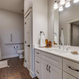 A modern bathroom with a double sink, large mirror, and sleek cabinetry, featuring bright lighting and a visible toilet area.