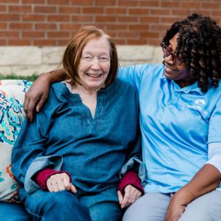 An elderly woman and her caregiver share a moment of laughter and companionship outdoors.