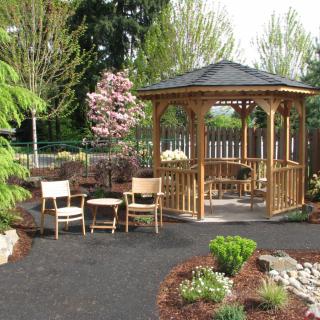 A tranquil garden setting featuring a wooden gazebo surrounded by lush greenery and chairs for relaxation.