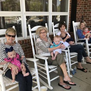 A group of women sitting on a porch in rocking chairs, each holding a baby.