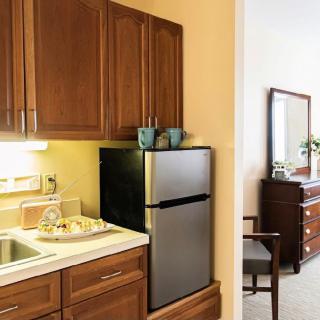 A cozy kitchenette with brown cabinets and a mini fridge next to a sink, leading into a room with a dresser.