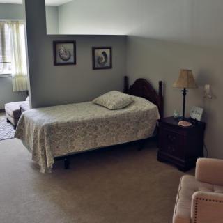A cozy bedroom with a single bed, nightstand, and lamp, featuring soft lighting from a nearby window.