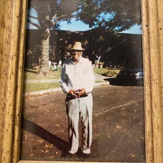 An elderly man stands outdoors, wearing a hat and casual attire.
