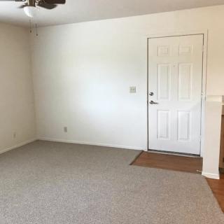An empty living room with a carpeted floor, featuring a ceiling fan and an entry door.