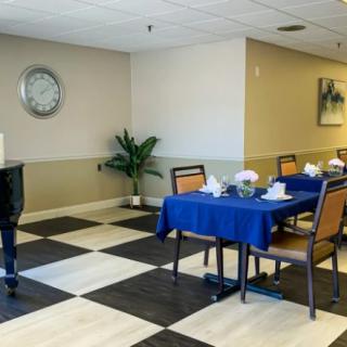 A cozy dining area with a checkered floor, featuring tables covered with blue tablecloths and a piano in the corner.