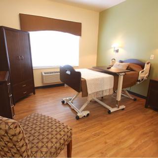A cozy nursing home bedroom with a hospital bed, armchair, and wooden floors.