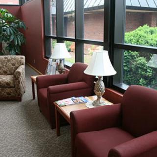 A cozy waiting area with red chairs, a lamp, and magazines by large windows overlooking a garden.