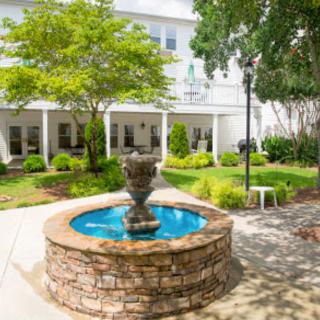 A serene garden area featuring a central fountain surrounded by lush greenery and a building in the background.