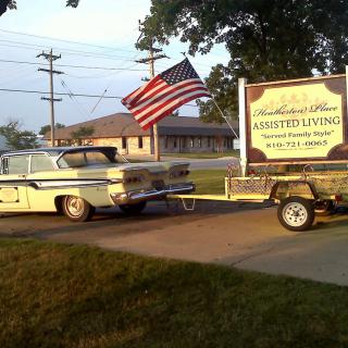 A vintage car towing a trailer with an American flag and signage in a suburban setting.