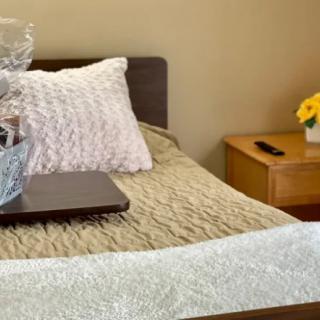 A cozy bedroom setting featuring a neatly made bed with accent pillows and a decorative gift basket on top. A nightstand with flowers completes the warm interior design.