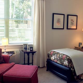 A cozy bedroom with a neatly made bed, artwork on the walls, and a comfortable chair by the window.