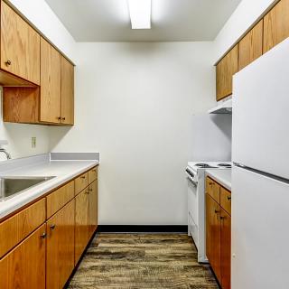 A narrow apartment kitchen with wood cabinets and white appliances.