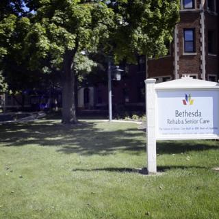 A healthcare facility sign stands on a grassy area in front of a building surrounded by trees.