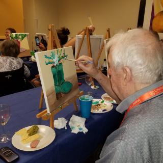 A senior man participating in an art class, painting on a canvas at an easel.