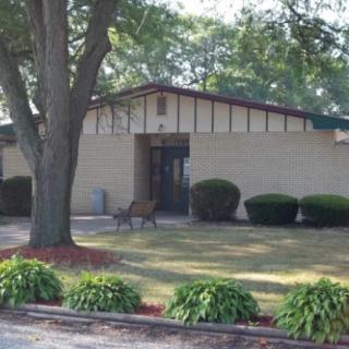 A small building with a tree and landscaped shrubs in the front yard.
