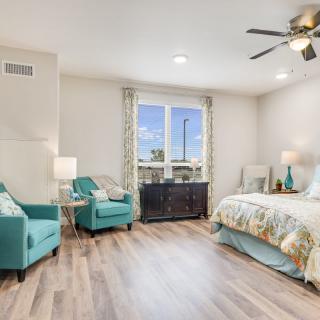 A modern and cozy bedroom featuring teal armchairs, a bed with floral bedding, and abundant natural light.