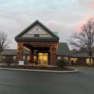 A brick building with a well-lit entrance surrounded by trees and a parking lot at dusk.