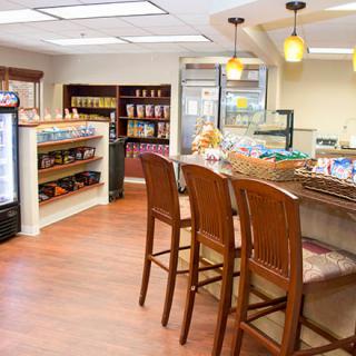 A cozy cafe interior featuring a seating area with high wooden chairs, a counter with baked goods, and shelves stocked with various items.