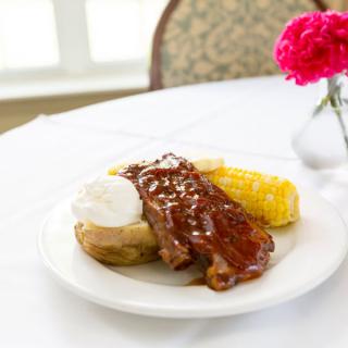 A delicious meal consisting of BBQ ribs, baked potato with sour cream, and corn on the cob served on a white plate.