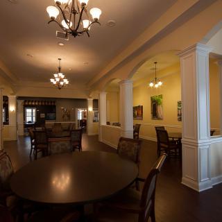 A spacious dining room illuminated by elegant chandeliers, featuring a large table surrounded by chairs and decorative columns.
