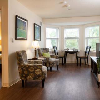 A cozy living room with comfortable chairs and a small table by the windows, featuring wooden flooring and wall artwork.