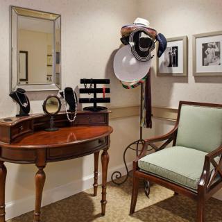 A cozy vintage corner featuring a wooden vanity desk, mirror, hat stand, and cushioned chair.