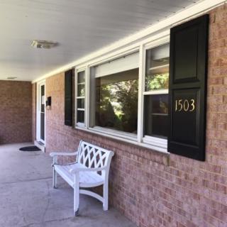 A brick house porch with a white bench and black shutters.