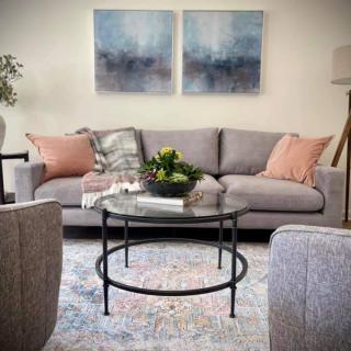 A cozy living room featuring a grey sofa with pink pillows, a round glass coffee table, and abstract artwork on the wall.
