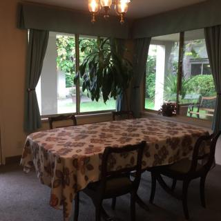 A cozy dining room with a table covered by a patterned cloth, surrounded by chairs and illuminated by a chandelier. Large windows allow daylight to brighten the space.