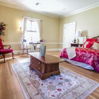 A cozy bedroom with a wooden floor, featuring a bed adorned with red bedding, a central rug, and elegant furnishings.