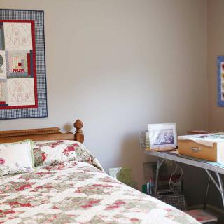 A cozy bedroom with a quilted bedspread and wall art, featuring a desk and lamp beside the bed.