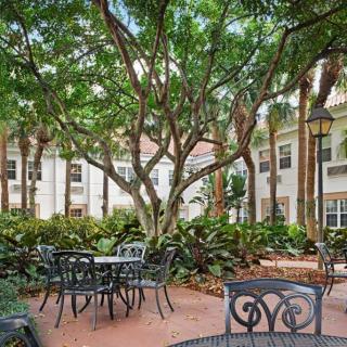 A serene outdoor patio area with tables and chairs surrounded by lush greenery and trees, adjacent to a building.