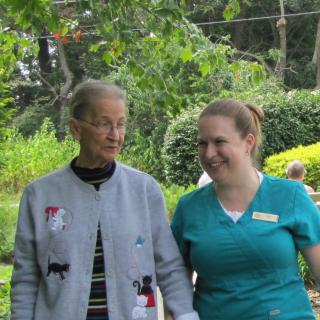 An elderly person walking with a caregiver in a park setting.