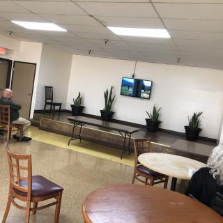 An empty waiting area with tables, chairs, a small stage with plants, and a television on the wall.