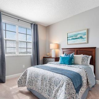 A well-decorated bedroom with a neatly made bed, featuring blue and white tones, a bedside lamp, and a framed painting above the headboard.