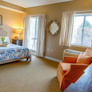 A cozy bedroom with a bed, armchair, and two large windows letting in natural light.