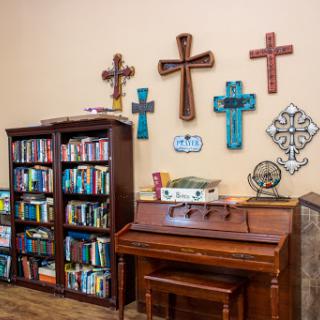 A cozy corner of a room featuring a wooden bookshelf filled with books, a piano, and an array of decorative crosses on the wall.