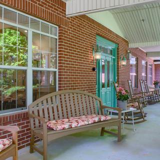 A cozy porch with wooden benches, rocking chairs, and decorative pillows against a brick wall.