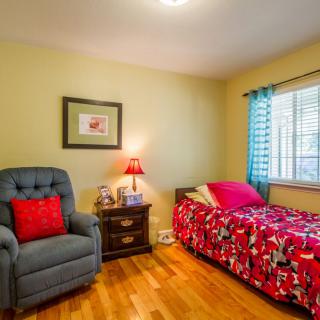A cozy bedroom featuring a single bed with red floral bedding, an armchair, nightstand with lamp, and a window with blue curtains.