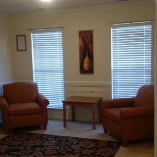 A cozy living room with two armchairs, a small table, and two windows with blinds.