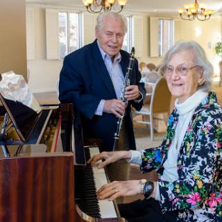 An elderly couple joyfully playing music together, with one on the piano and the other on the clarinet.