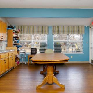 A bright dining room with blue walls, featuring a wooden table and kitchen cabinets, illuminated by natural light through large windows.