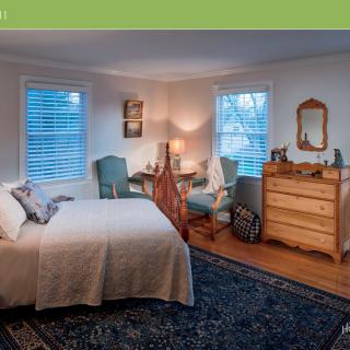 A cozy and well-decorated bedroom featuring a bed, chairs, dresser, and rug with natural light from windows.