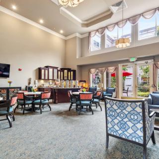 A spacious and well-lit dining area with elegant decor, featuring multiple tables and chairs, large windows, and a chandelier.
