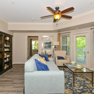 A cozy living room featuring a light blue sofa, armchair, bookshelf, and glass coffee table with a view of the outdoor greenery through large glass doors.