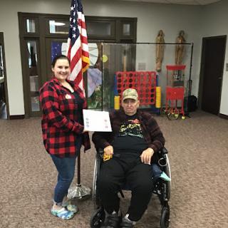 A young woman stands beside an elderly person in a wheelchair, proudly holding a certificate next to an American flag.