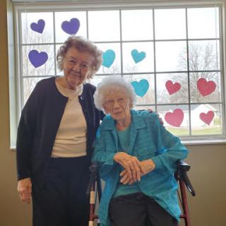 Two joyful women pose together in front of a window decorated with colorful hearts.