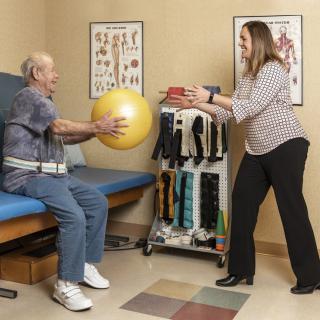 A therapist assists an elderly patient with exercises using an exercise ball in a healthcare setting.