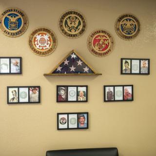 A wall display featuring military seals, an American flag in a triangular case, and several framed pictures.